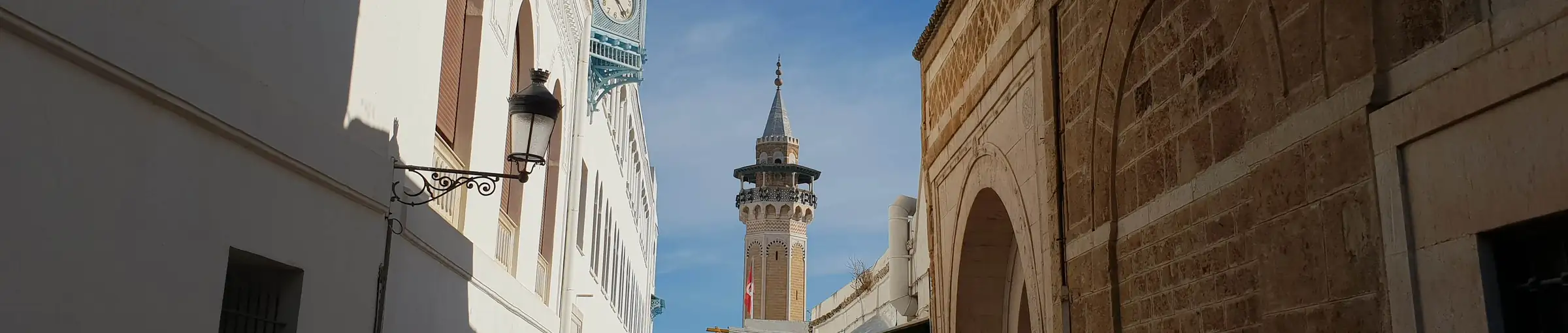 Ruelle de la médina de Tunis entre deux façades, avec la tour de l’horloge et le minaret d’une mosquée au fond